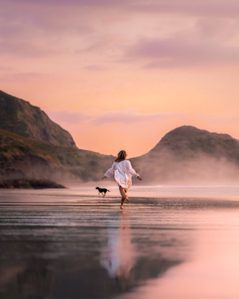 Back view full length slim female in white males shirt running with dog on scenic wet seacoast against rough cliffs on early evening